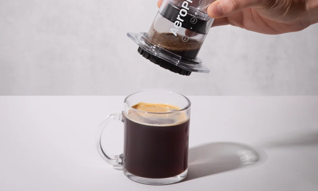 A person removes an AeroPress Clear from the top of a mug of brewed coffee.